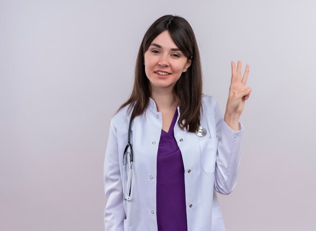 Przekonana młoda kobieta lekarz w szlafroku medycznym z stetoskopem gesty trzy palcami na odosobnionym białym tle z miejsca na kopię