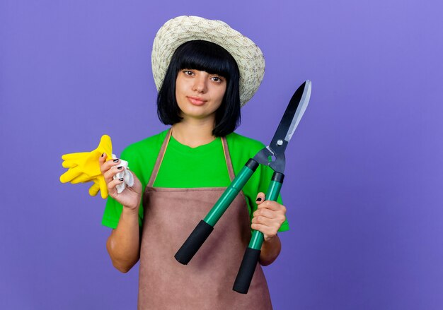 Przekonana, młoda kobieta ogrodnik w mundurze na sobie kapelusz ogrodniczy posiada maszynki do strzyżenia i rękawiczki