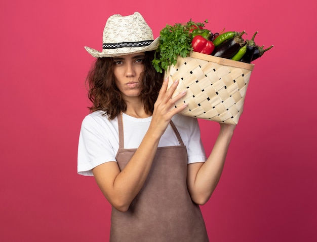 Przekonana, młoda kobieta ogrodnik w mundurze na sobie kapelusz ogrodniczy stawiając kosz warzyw na ramieniu