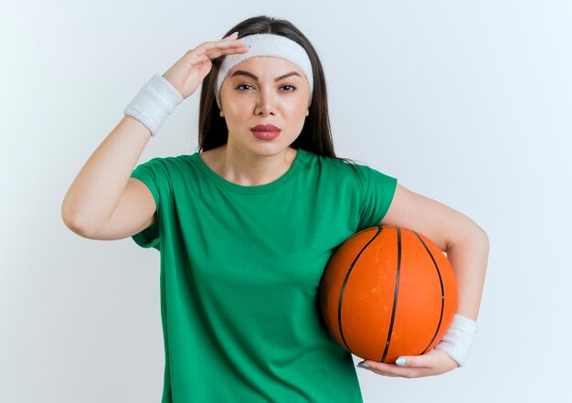 Przekonana, młoda kobieta sportowy, noszenie opaski i opaski na rękę, trzymając piłkę do koszykówki, patrząc w dal