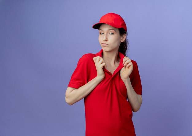 Przekonana, młoda ładna dziewczyna dostawy ubrana w czerwony mundur i czapkę, chwytając kołnierz jej koszulki na białym tle na fioletowym tle z miejsca na kopię