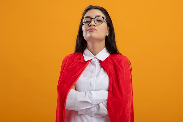 Przekonana, młoda superwoman w okularach stojących z zamkniętą postawą, patrząc z przodu na białym tle na pomarańczowej ścianie