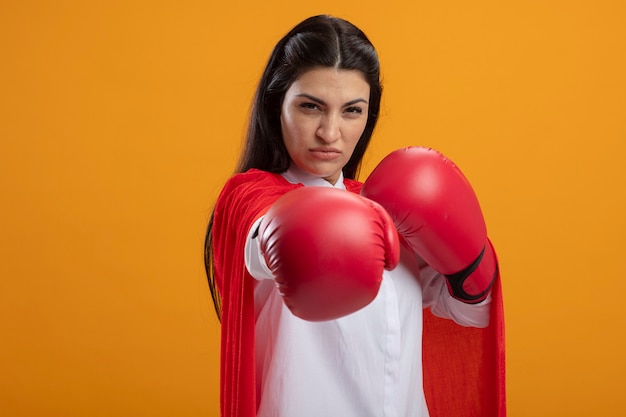 Przekonana, młoda superwoman w rękawiczkach pudełkowych, patrząc na przód, robi gest bokserski na białym tle na pomarańczowej ścianie