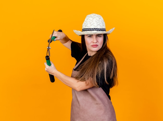 Przekonana, piękna dziewczyna ogrodnik na sobie kapelusz mundurowy i ogrodnictwo trzymając maszynki do strzyżenia na pomarańczowo