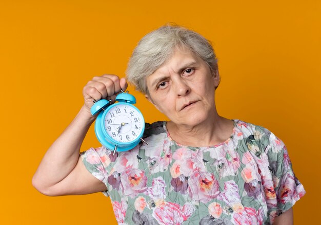Przekonana, starsza kobieta trzyma budzik na białym tle na pomarańczowej ścianie