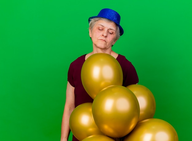 Przekonana, starsza kobieta ubrana w kapelusz partii stoi i patrzy na balony z helem na białym tle na zielonej ścianie z miejsca na kopię