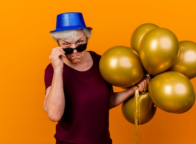 Przekonana, starsza kobieta w kapeluszu partii trzyma okulary przeciwsłoneczne i balony z helem na białym tle na pomarańczowej ścianie z miejsca na kopię