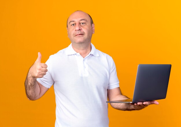 Przekonany, dorywczo dojrzały mężczyzna trzyma laptopa jego kciuk w górę na białym tle na żółtym tle