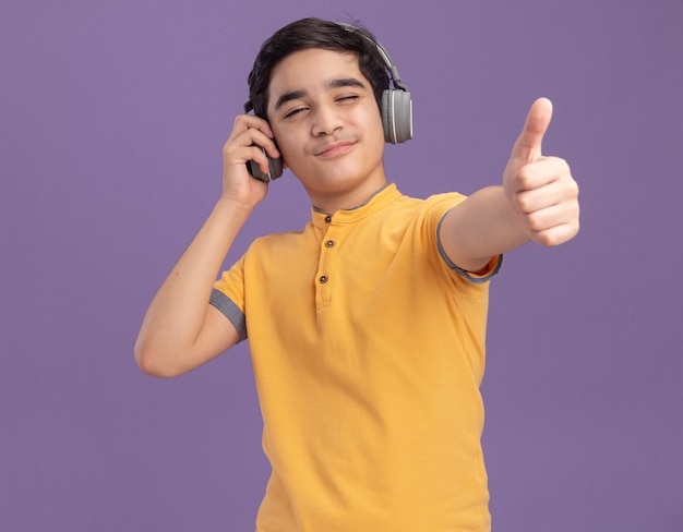 Przekonany, młody chłopiec kaukaski nosi i chwyta słuchawki, mrugając pokazując kciuk na białym tle na fioletowej ścianie