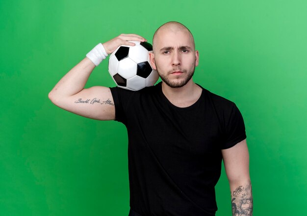 Przekonany, młody człowiek sportowy noszenia opaski na rękę, kładąc piłkę na ramieniu na białym tle na zielonej ścianie