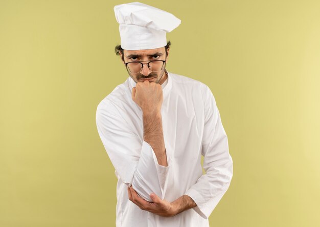 Przekonany, młody mężczyzna kucharz na sobie mundur szefa kuchni i okulary, kładąc rękę na brodzie