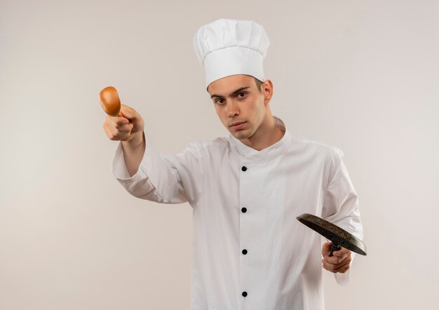 Przekonany, młody mężczyzna kucharz ubrany w mundur szefa kuchni, trzymając patelnię i wyciągając łyżkę na odizolowanej białej ścianie