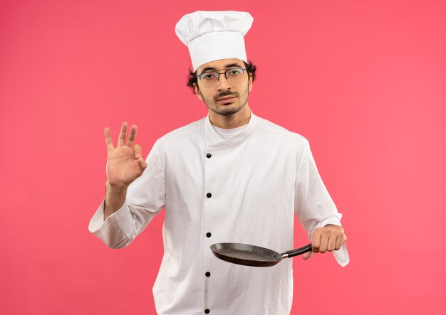 Przekonany, młody mężczyzna kucharz w mundurze szefa kuchni i okularach, trzymając patelnię i pokazując gest okey