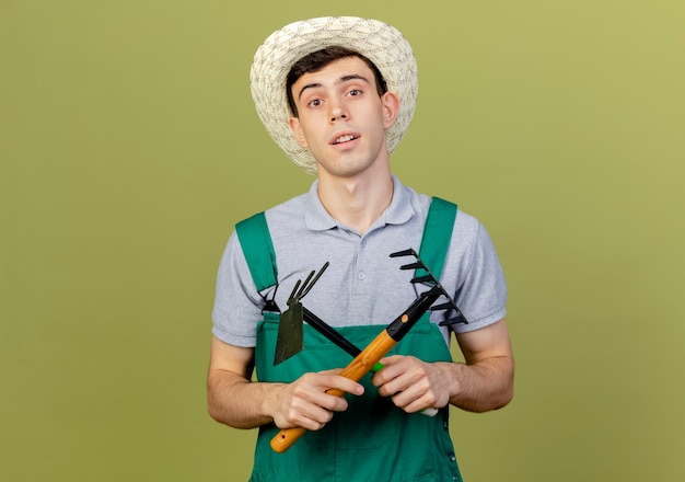 Przekonany, młody mężczyzna ogrodnik w kapeluszu ogrodniczym krzyżuje grabie i motyka