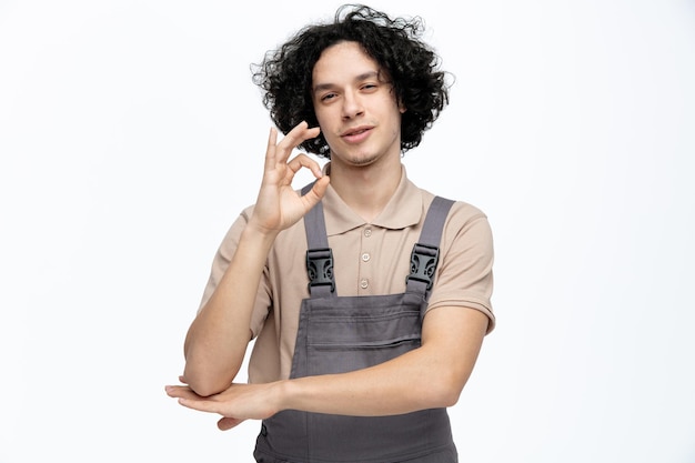 Przekonany, młody mężczyzna pracownik budowlany ubrany w mundur, patrząc na kamery pokazujący znak porządku, trzymając rękę pod łokciem na białym tle