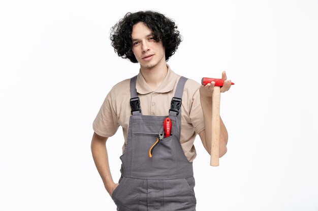 Przekonany, młody mężczyzna pracownik budowlany ubrany w mundur, trzymając rękę w kieszeni trzymając młotek patrząc na kamery z śrubokrętem i szczypcami w kieszeni na białym tle