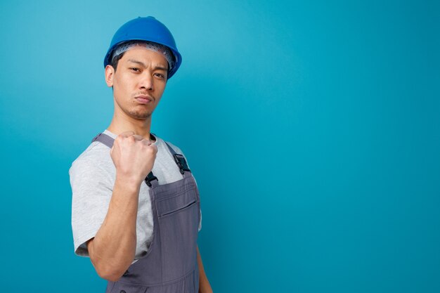 Przekonany, młody pracownik budowlany na sobie hełm ochronny i mundur stojący w widoku profilu robi tak gest