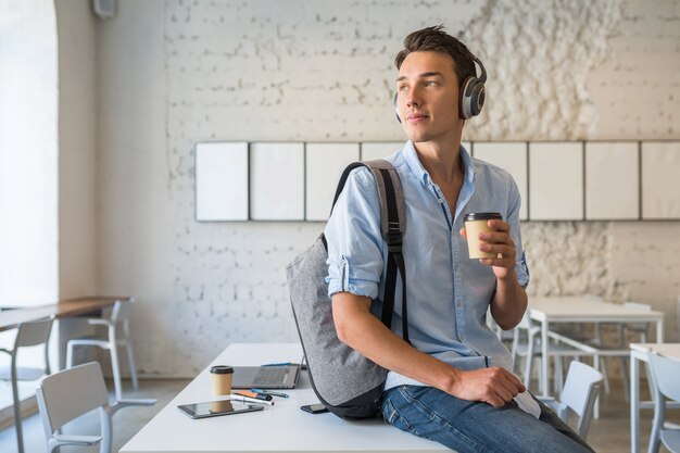 Przekonany, młody przystojny mężczyzna siedzi na stole w słuchawkach z plecakiem w biurze współpracującym, pije kawę