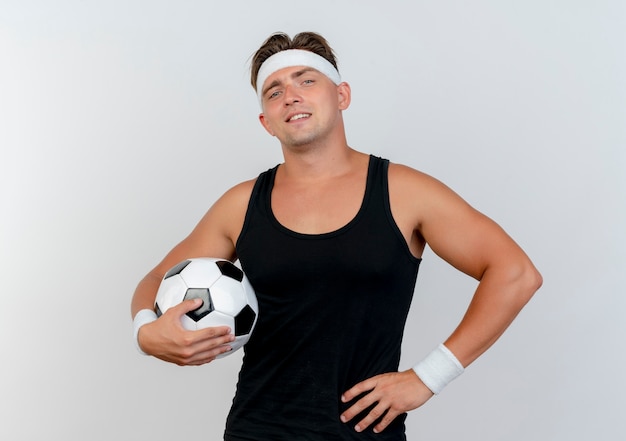 Przekonany, młody przystojny mężczyzna sportowy, noszenie opaski i opaski na rękę, trzymając piłkę nożną i kładąc rękę na talii na białym tle