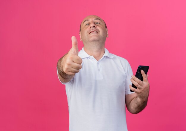Przekonany, przypadkowy dojrzały biznesmen, trzymając telefon komórkowy i pokazując kciuk do góry na białym tle na różowym tle z miejsca na kopię