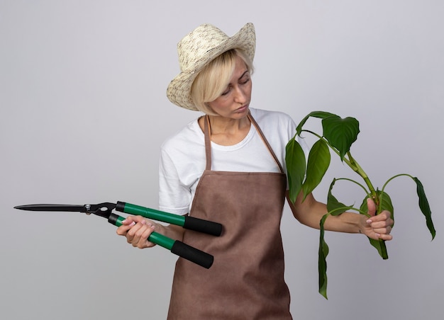 Przemyślana blond ogrodniczka w średnim wieku w mundurze, w kapeluszu, trzymająca nożyce do żywopłotu i roślin, patrząc na roślinę