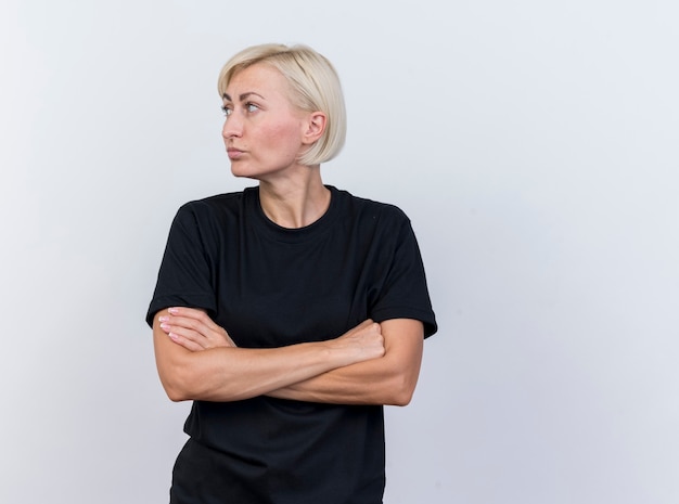 Przemyślana blondynka w średnim wieku kobieta stojąca z zamkniętą posturą, patrząc z boku na białym tle na białej ścianie