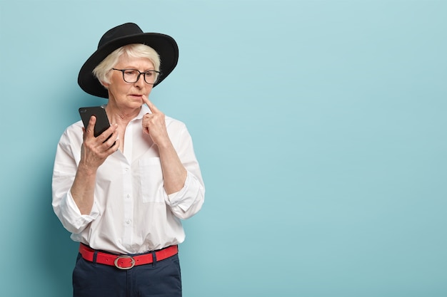Przemyślana, charyzmatyczna seniorka ubrana w modny strój, myśli o treści wiadomości, trzyma w ręku nowoczesny telefon komórkowy