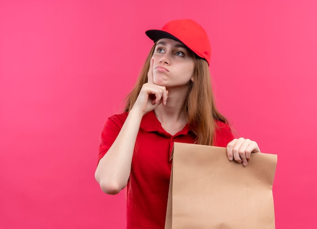 Bezpłatne zdjęcie przemyślana młoda dziewczyna dostawy w czerwonym mundurze, trzymając papierową torbę i kładąc rękę pod brodą na odizolowanej różowej ścianie z miejscem na kopię