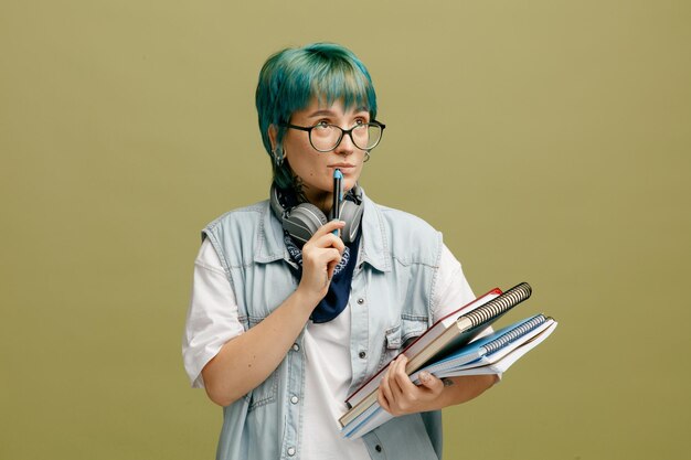 Przemyślana Młoda Studentka W Okularach Bandana I Słuchawki Na Szyi Trzymająca Notesy Patrząc W Górę Dotykając Wargi Piórem Na Białym Tle Na Oliwkowo-zielonym Tle