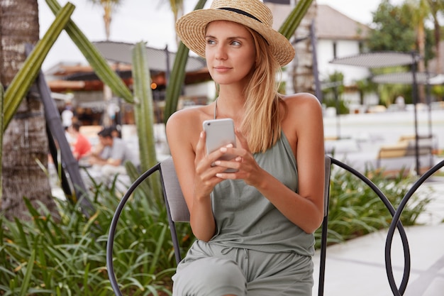 Przemyślana piękna młoda kobieta myśli o czymś, odtwarzając coś w kawiarni na świeżym powietrzu ze smartfonem w letni dzień, odwraca wzrok, podłączona do bezprzewodowego internetu do komunikacji online