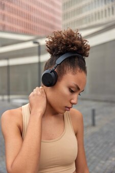 Przemyślana zmęczona kobieta z kręconymi włosami trzyma rękę na szyi skupiona w dół słucha muzyki w bezprzewodowych słuchawkach nosi sportowy stanik ma regularne poranne pozy treningowe na zewnątrz na rozmytym tle.
