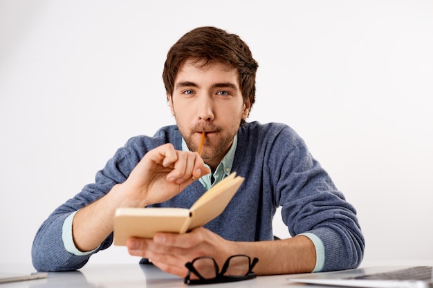 Przemyślany, kreatywny męski dziennikarz lub pisarz, gryzący ołówek, trzymający notatnik, harmonogram pisania, wyglądający na myślącego