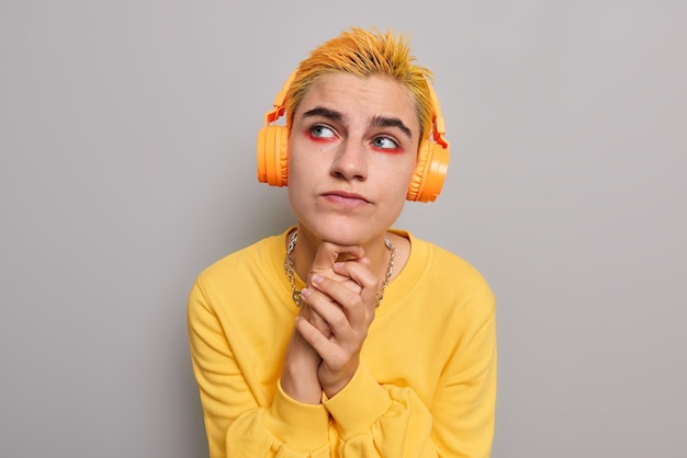 Bezpłatne zdjęcie przemyślany meloman o niecodziennym wyglądzie myśli o czymś słuchając muzyki w słuchawkach, trzymając ręce pod brodą, nosi jaskrawy makijaż na tle szarości. koncepcja młodzieży.