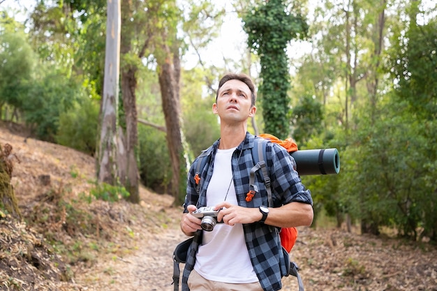 Przemyślany mężczyzna trzyma aparat, odwraca wzrok i stoi na drodze. Kaukaski turysta eksplorujący przyrodę i fotografujący przyrodę. Koncepcja turystyki, przygody i wakacji letnich