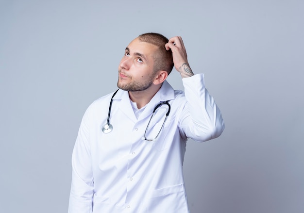 Przemyślany Młody Lekarz Płci Męskiej Ubrany W Szlafrok Medyczny I Stetoskop Na Szyi, Patrząc W Górę I Drapiąc Się Po Głowie Na Białej ścianie