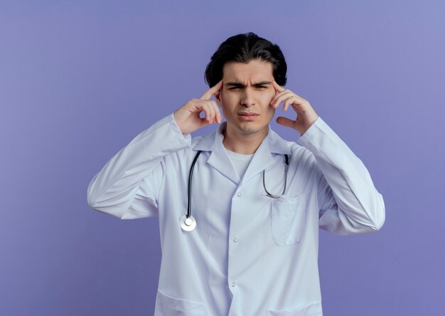 Przemyślany młody lekarz płci męskiej ubrany w szlafrok medyczny i stetoskop patrząc na bok, wskazując palcami na głowę odizolowaną na fioletowej ścianie z miejscem na kopię