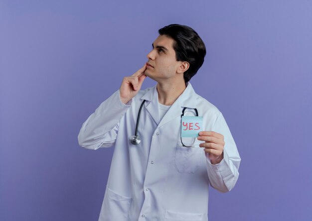 Przemyślany młody lekarz płci męskiej ubrany w szlafrok medyczny i stetoskop pokazujący tak notatkę patrząc na stronę dotykającą brody odizolowaną na fioletowej ścianie z miejscem na kopię