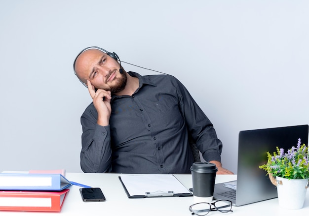 Przemyślany Młody łysy Mężczyzna Call Center Sobie Zestaw Słuchawkowy Siedzi Przy Biurku Z Narzędzi Pracy Kładąc Palec Na świątyni Patrząc W Górę Na Białym Tle Na Białej ścianie