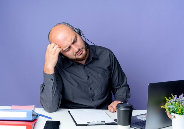 Przemyślany młody łysy mężczyzna call center sobie zestaw słuchawkowy siedzi przy biurku z narzędziami pracy kładąc rękę na głowie patrząc w dół na białym tle na fioletowej ścianie