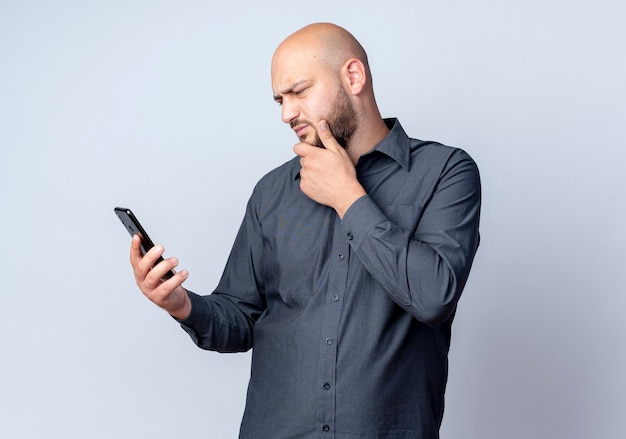 Przemyślany młody łysy mężczyzna call center trzymając i patrząc na telefon komórkowy z ręką na brodzie na białym tle na białej ścianie
