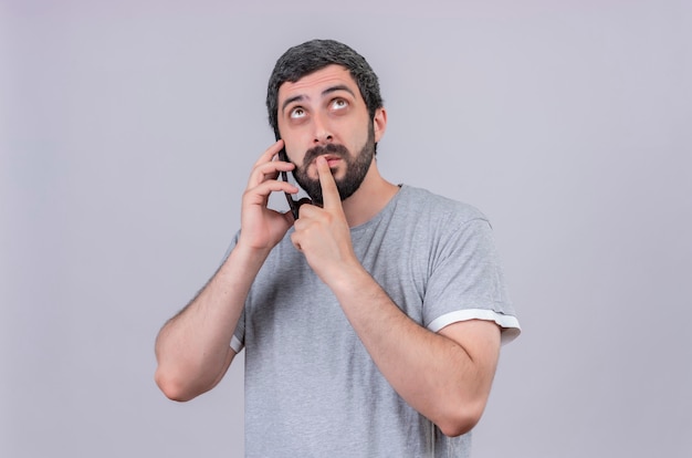 Przemyślany młody przystojny mężczyzna rozmawia przez telefon kładąc palec na ustach i patrząc w górę na białym tle na białej ścianie
