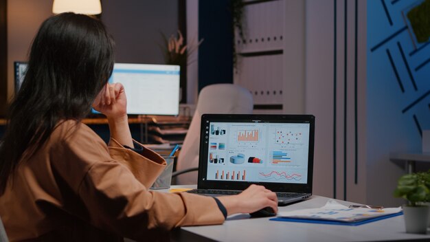 Przepracowana bizneswoman pisania statystyk finansowych na laptopie, siedząca przy biurku w biurze firmy startowej późno w nocy