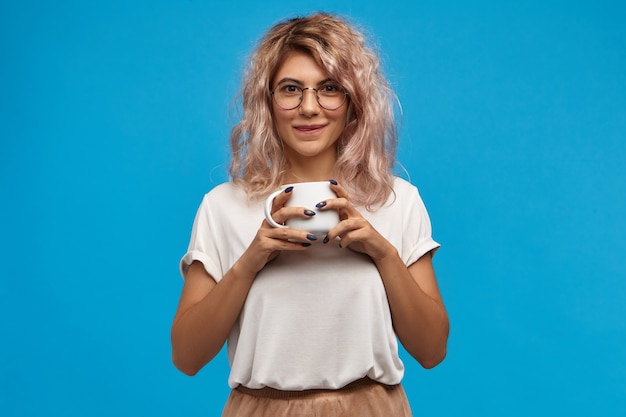 Bezpłatne zdjęcie przepyszny. portret młodej kobiety ładny nerdy na sobie okrągłe okulary, ciesząc się słodką gorącą czekoladą. ładna dziewczyna z różowawymi włosami, trzymając biały kubek, pijąc dobrą świeżą kawę