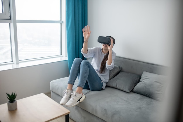 Przerażona młoda kobieta w goglach VR, siedząca na kanapie w salonie
