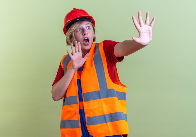Przerażona młoda konstruktorka w mundurze trzymająca ręce z boku odizolowana na oliwkowozielonej ścianie