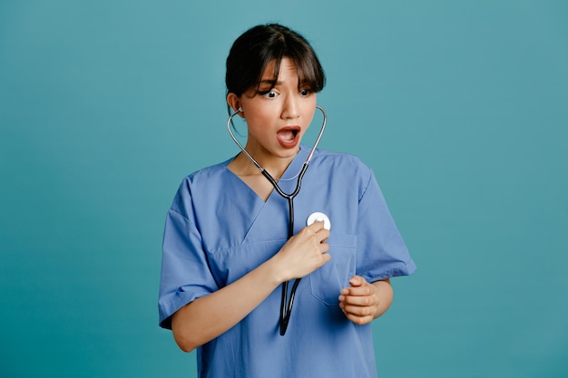 Przerażona młoda lekarka nosząca jednolity stetoskop piąty na białym tle na niebieskim tle
