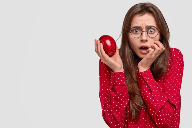 Przerażona piękna emocjonalna dama wygląda bezpośrednio ze strasznym wyrazem, trzyma w dłoni świeże czerwone jabłko