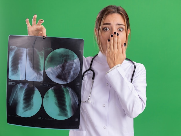 Bezpłatne zdjęcie przestraszona młoda lekarka nosząca szatę medyczną ze stetoskopem trzymająca prześwietlenie i zakryte usta ręką odizolowaną na zielonej ścianie