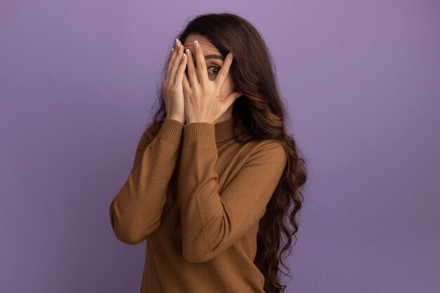 Przestraszona młoda piękna dziewczyna ubrana w brązowy sweter z golfem zakryła twarz rękami odizolowanymi na fioletowej ścianie