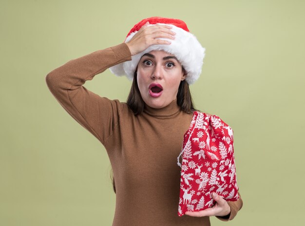 Przestraszona młoda piękna dziewczyna w kapeluszu Boże Narodzenie trzymając worek Boże Narodzenie kładąc rękę na głowie na białym tle na oliwkowym tle